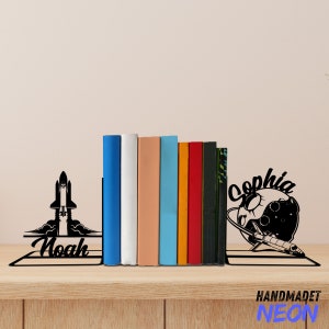 Puede incluir: Dos sujetalibros de metal negro con un cohete y una luna creciente con estrellas. El sujetalibros del cohete tiene el nombre "Noah" y el sujetalibros de la luna tiene el nombre "Sophia". Los sujetalibros sostienen una fila de libros coloridos.