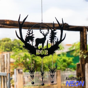 May include: Black metal deer silhouette wind chime with a deer, trees, and the name "BOB". The wind chime has three hanging deer heads with bells. The design is attached to a chain for hanging.