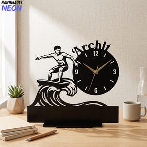 May include: Black decorative clock featuring a surfer riding a wave. The clock face is round with gold hands and the name "Archit" above. The base is rectangular. The clock is on a wooden surface.