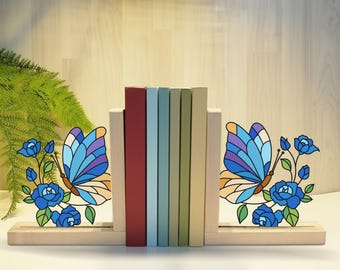 May include: Wooden bookends with a butterfly and rose design. The bookends feature a light wood base and support, with a colorful butterfly and blue rose motif. A row of books in various colors sits between the bookends.
