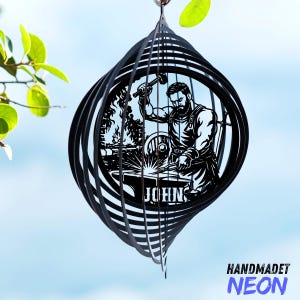 Pode incluir: Moinho de vento de metal preto com um design de ferreiro. A obra de arte retrata um ferreiro no trabalho, com o nome "JOHN" na parte inferior. O moinho de vento tem um design em espiral e o texto "HANDMADET NEON" na parte inferior.