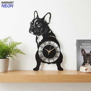 May include: Black metal wall clock in the shape of a French Bulldog. The clock face is round with Roman numerals and gold-colored hands. The clock is mounted on a white wall above a wooden shelf. The text "HANDMADET NEON" is in the upper left corner.
