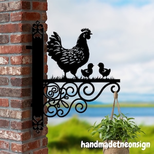 May include: Black metal silhouette of a hen and chicks, mounted on a decorative scrollwork bracket. The bracket is attached to a brick wall. A hanging plant is visible in the foreground.