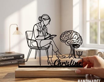 May include: A black metal wire art sculpture featuring a person sitting in a chair writing on a notepad, next to a brain illustration and the name Christine. The sculpture is mounted on a rectangular light wood base.
