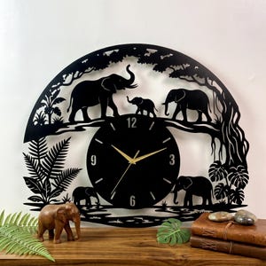 May include: Black metal wall clock featuring a silhouette of elephants in a jungle scene. The clock face is black with gold hands. A wooden elephant figurine sits on a wooden surface.