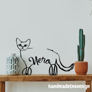 May include: Black metal wall art of a cat with the name "Vera" written in cursive. The art is displayed on a wooden shelf with a patterned vase and a potted cactus.