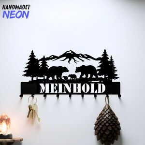 May include: Black metal key holder featuring a mountain range, trees, and bear family silhouette. The name "MEINHOLD" is displayed below the silhouette. Includes hooks for keys and a decorative pinecone.