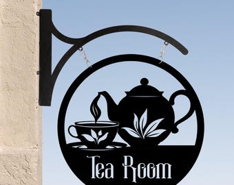 May include: A black circular metal sign featuring a cutout silhouette of a teapot pouring into a teacup. The text Tea Room is displayed in white lettering at the bottom. The sign is attached to a black metal bracket for wall mounting.
