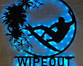 May include: Black metal silhouette of a surfer riding a wave with palm trees and hibiscus flowers. The silhouette is illuminated with blue LED lights and the words "WIPEOUT" are displayed below.