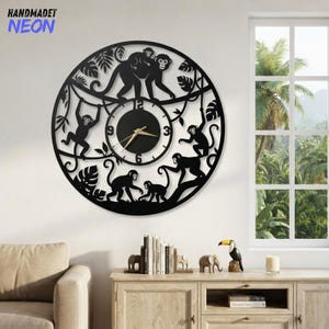 Puede incluir: Un reloj de pared circular negro con un diseño recortado detallado de varios monos trepando entre lianas y hojas de la selva. La esfera del reloj está centrada con un fondo negro, números blancos y manecillas de color dorado.