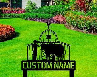 May include: A black metal garden sign featuring a silhouette of a barn with a rooster weather vane on top, a person wearing a hat, and a cow. The sign includes a rectangular base with the text CUSTOM NAME in bold, block capital letters.