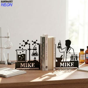 Op de afbeelding: Een paar boekensteunen van zwart metaal met wetenschappelijke silhouetten. De ene kant toont een bekerglas, een kolf boven een vlam en moleculaire structuren. De andere kant toont een wetenschapper met een kolf naast een vijzel en reageerbuizen. Beide stukken zijn voorzien van de naam MIKE.