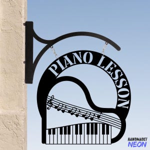 Pode incluir: Placa de metal preto com uma silhueta de piano, notas musicais e o texto "PIANO LESSON" em um círculo. A placa está pendurada em um suporte de metal preto.