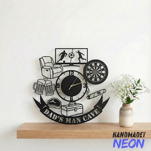 May include: Black metal wall clock with a "Dad's Man Cave" theme. The clock features a football game on TV, a dartboard, beer mugs, a cigar, and a toolbox. The clock is on a wooden shelf with a vase of flowers.