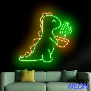 Könnte beinhalten: Ein Neonschild mit einem grünen Dinosaurier, der einen Kaktus in einem Topf hält. Der Dinosaurier hat eine orangefarbene Umrandung und befindet sich vor einem schwarzen Hintergrund. Der Text "HANDMADET NEON" ist sichtbar. Das Schild befindet sich über einem grünen Sofa mit Kissen.