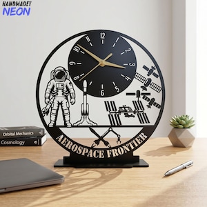 May include: Black laser-cut desk clock with an aerospace theme. The clock features an astronaut, rocket, and space station silhouettes. The clock face is black with gold hands and the words "AEROSPACE FRONTIER". The clock is approximately 10 inches tall.