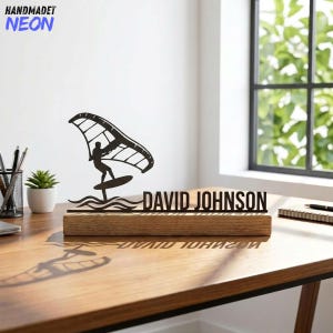 May include: A black laser-cut desk sign featuring a windsurfer design and the name "DAVID JOHNSON". The sign is mounted on a rectangular wooden base. The words "HANDMADET NEON" are in the upper left corner.