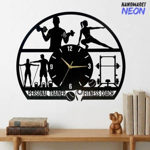 May include: Black metal wall clock featuring silhouettes of people exercising, dumbbells, and gym equipment. The clock face has gold hands and the words "PERSONAL TRAINER" and "FITNESS COACH". The text "HANDMADET NEON" is in the upper right corner.