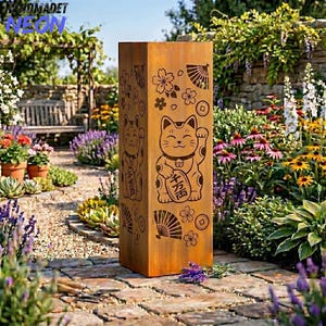 Puede incluir: Una escultura de jardín alta y rectangular de metal envejecido. La escultura presenta diseños grabados de un gato que llama, flores de cerezo y abanicos. La escultura está situada en un jardín con un camino de ladrillos y varias flores.