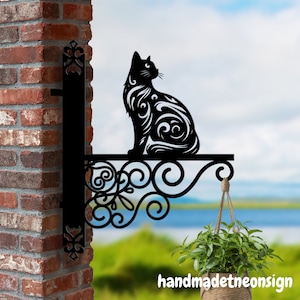 May include: Black metal wall-mounted sign featuring a stylized cat design with intricate swirls. The sign has a decorative scrollwork design and a hanging hook. The sign is mounted on a brick wall with a green plant hanging below.