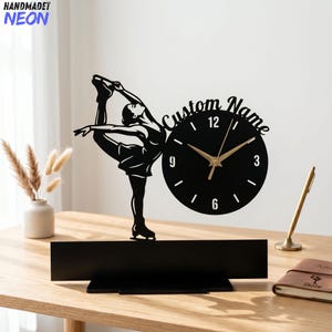 May include: Black decorative clock featuring a silhouette of a figure skater in a graceful pose. The clock has a round face with gold hands and the text "Custom Name". The clock is mounted on a black base. The word "HANDMADET NEON" is in the top left corner.