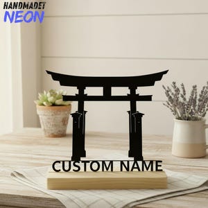 Könnte beinhalten: Schwarze Metall-Torii-Tor-Silhouette auf einem Holzsockel, mit dem Text "CUSTOM NAME". In der oberen linken Ecke steht der Text "HANDMADET NEON" in Blau. Das Tor ist ein dekoratives Element.