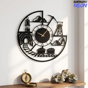 May include: A circular black metal wall clock featuring a mining-themed silhouette design. The clock face has gold-coloured hands and displays a tunnel, train tracks, a lantern, a pickaxe, a headframe, and two mine carts. The text HANDMADET NEON is visible.