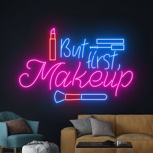 Puede incluir: Un letrero de neón con luces rosas y azules que dice "But first, makeup". El letrero incluye un lápiz labial y una brocha de maquillaje.