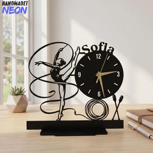 May include: A black tabletop clock featuring a silhouette of a rhythmic gymnast with a ribbon, the name Sofia, a circular clock face with gold hands, and a pair of clubs. The design is mounted on a rectangular base, creating a decorative piece.
