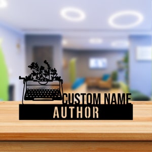 May include: Black metal sign featuring a silhouette of a vintage typewriter with flowers on top. The sign reads "CUSTOM NAME AUTHOR" in a stencil font.