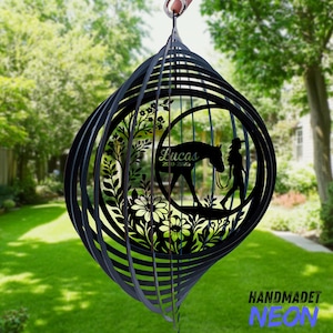 May include: Black metal wind spinner featuring a horse and rider silhouette within a floral design. The name "Lucas" and dates are displayed. The spinner is suspended outdoors with a green lawn and trees in the background.
