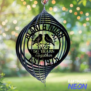 May include: Black metal wind spinner with a spiral design. The center features a circular design with two birds, the names "Blani & Noah", and the text "50 Years Together Since 1974". The bottom right corner has the text "HANDMADET NEON".