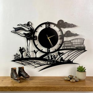May include: Black metal wall clock featuring a figure skater, ice skating silhouettes, and a figure skating pavilion. The clock has a round face with gold hands. Two brown ice skates are on a wooden shelf below the clock.