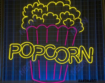 May include: A neon sign with the word "POPCORN" in yellow letters and a pink and blue popcorn bucket with yellow popcorn on top. The sign is handmade and says "HANDMADET NEON" at the bottom.