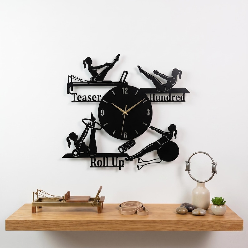 May include: Black metal Pilates-themed wall clock featuring silhouettes of women performing exercises. The clock face is round with gold hands and numbers. The words "Teaser," "Hundred," and "Roll Up" are included in the design.