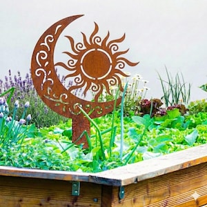 May include: A decorative metal garden stake with a sun and crescent moon design. The sun has stylized rays and the moon has decorative cutouts. The stake is brown and is placed in a garden bed with green plants and purple flowers.