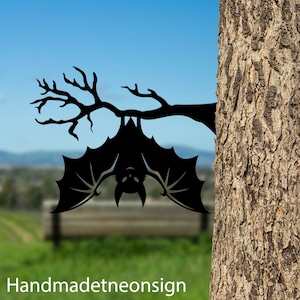 May include: Black metal silhouette of a bat hanging upside down from a tree branch. The bat is in a classic Halloween style. The background is blurred and out of focus.