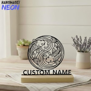 May include: Black metal koi fish design with the text "CUSTOM NAME" on a wooden base. The design features two fish in a circular pattern, with the words "HANDMADET NEON" in blue.