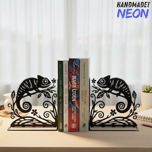 May include: Black metal bookends featuring a chameleon design. The bookends have a decorative floral and leaf pattern. The text "HANDMADET NEON" is visible in the upper right corner. Books are placed between the bookends.
