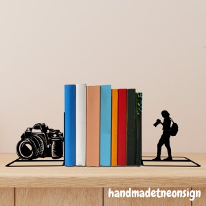 May include: Black metal bookends shaped like a camera and a person holding a camera. The bookends are holding up a row of colorful books.