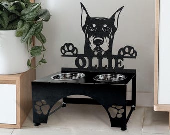May include: Black metal dog bowl stand with two bowls. The stand features a silhouette of a Doberman Pinscher with the name "Ollie" underneath. The stand has paw prints on the sides.