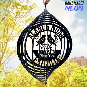 Puede incluir: Un molinillo de viento de metal negro con un dise&ntilde;o circular que presenta dos p&aacute;jaros posados en una rama. Se muestra el texto "ALAN & NOAH" y "10 YEARS Together EST 2013". Las palabras "HANDMADET NEON" est&aacute;n en la esquina superior derecha.