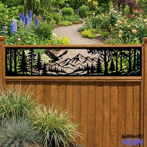 May include: A rectangular black metal decorative panel featuring a laser-cut silhouette design of a soaring eagle, a mountain range, and a dense forest of pine trees. The panel is mounted horizontally atop a vertical wooden fence section.