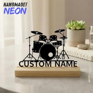 May include: A black metal drum kit silhouette with cymbals and stands, mounted on a wooden base. The words "CUSTOM NAME" are cut out below the drum kit. The words "HANDMADET NEON" are in blue above the drum kit.