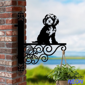 Puede incluir: Macetero de pared de metal negro con un diseño decorativo de volutas. Una pequeña silueta de perro se sienta en la parte superior del macetero. El macetero está sujeto a una pared de ladrillo y tiene una planta colgante delante.