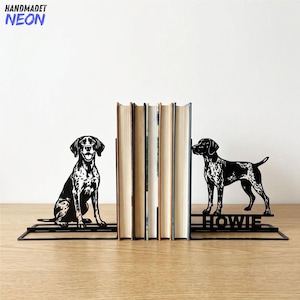 May include: A pair of black metal bookends featuring a silhouette of a spotted dog. One bookend shows the dog sitting, while the other shows the dog standing with the name HOWIE cut out in the base. The bookends support a row of five books.