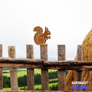 May include: A brown metal squirrel silhouette perched on a wooden fence post. The rustic fence has horizontal and vertical wooden beams. The background features a green field, trees, and a cloudy sky. The image has a rural, outdoorsy theme.