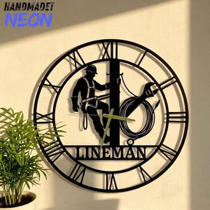 May include: A black circular wall clock featuring Roman numerals and a central silhouette of a lineman working on a utility pole. The design includes a wrench and coiled wire, with the word LINEMAN displayed prominently at the bottom center.