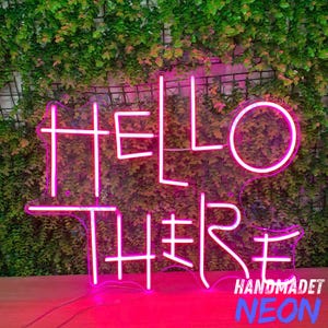 Puede incluir: Un letrero de neón rosa brillante que dice "HELLO THERE" en una fuente estilizada. El letrero está sobre un fondo de follaje verde y una superficie de madera. Las palabras "HANDMADET NEON" están escritas en blanco y azul.