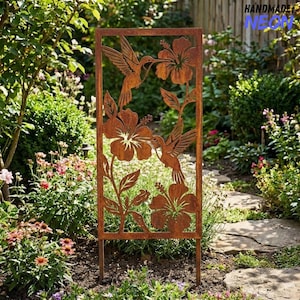May include: A rectangular, rust-coloured metal garden screen with cut-out designs of hummingbirds and hibiscus flowers. The screen has a frame and two stakes for ground placement. The words "HANDMADE" and "NEON" are visible in the top right corner.